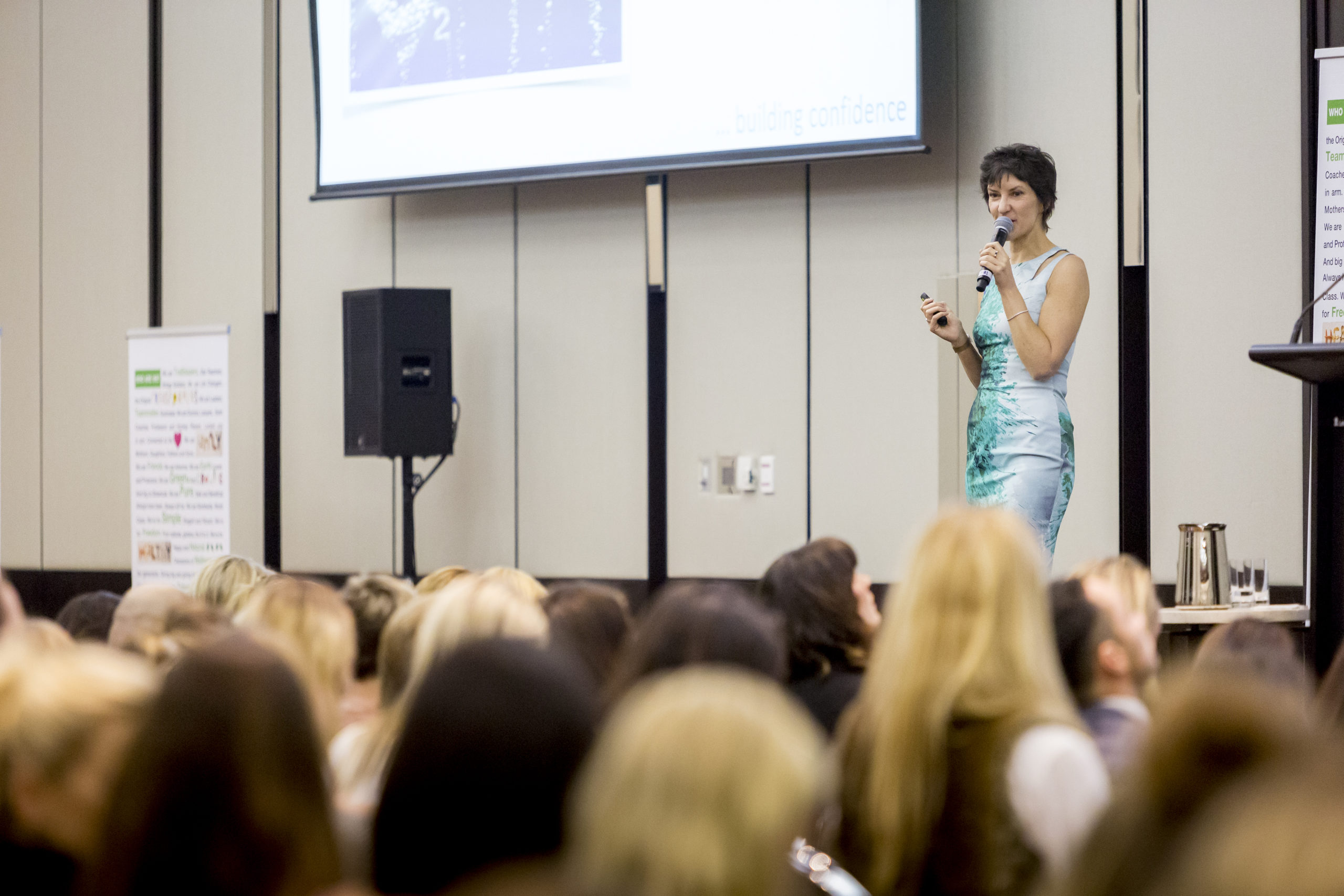 Sarah Denholm presenting on a large stage holding a microphone with screen behind her
