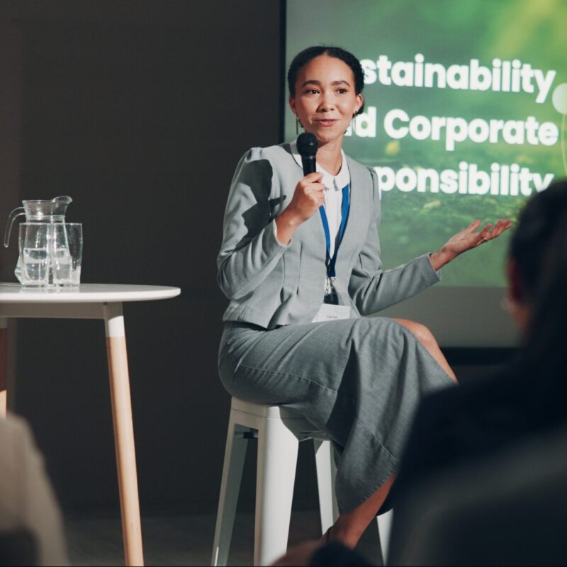 woman seated with microphone speaking on stage discussing sustainability and corporate governance, named on the screen behind her.