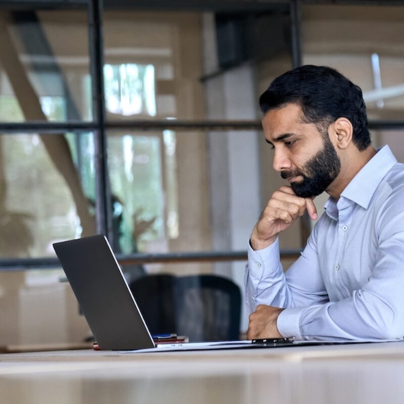 Thoughtful concentrated indian business man entrepreneur investor manager using computer, watching webinar working in office analyzing online data market thinking doing web research looking at laptop.